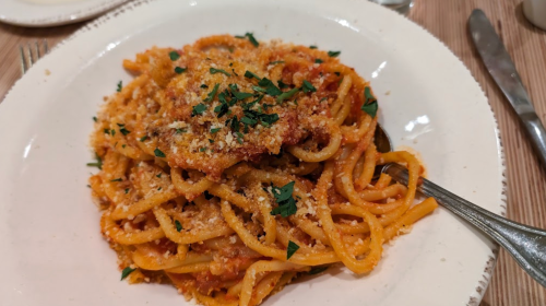 A plate of spaghetti topped with breadcrumbs and parsley, served on a white dish with a fork beside it.